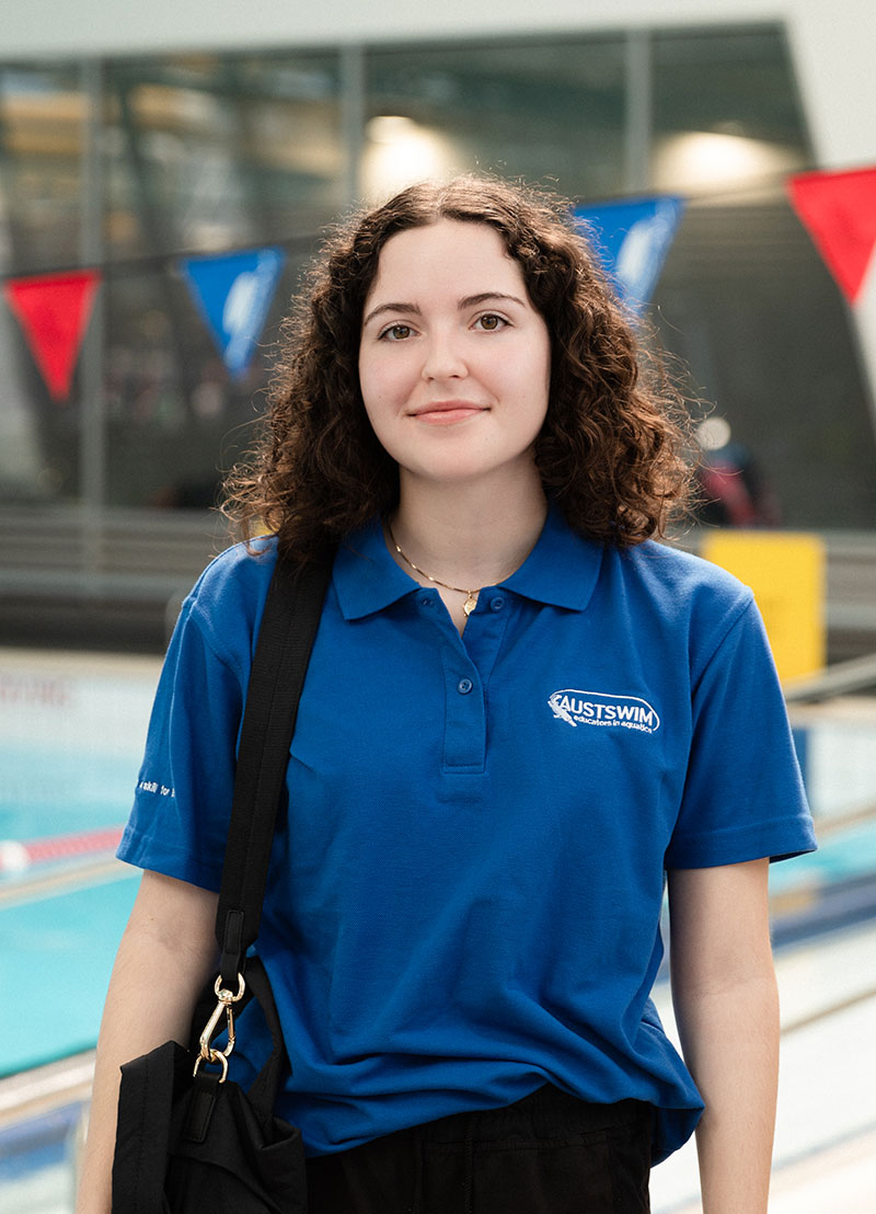 Image of swim teacher wearing a royal blue AUSTSWIM polo shirt. Smiling at the camera standing next to a pool with a bag over her shoulder.