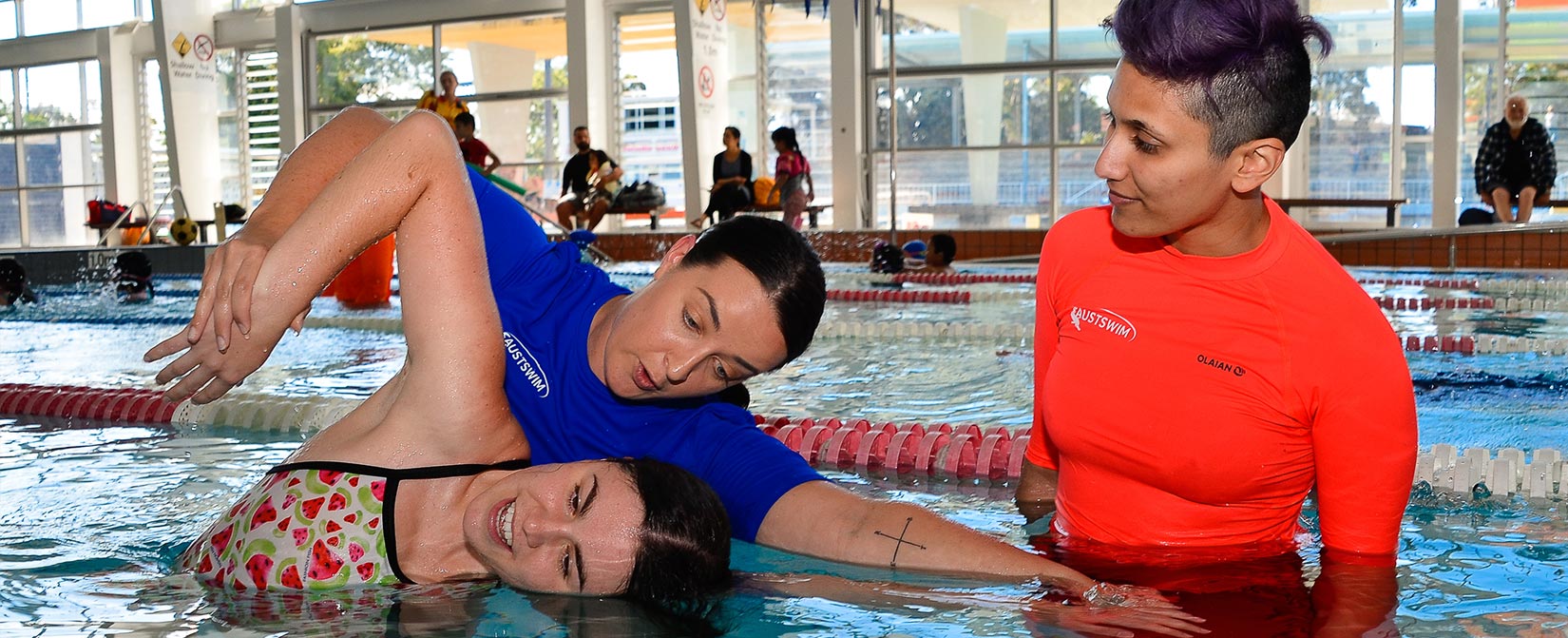 AUSTSWIM teacher and teacher in training teach a child in a swimming pool.